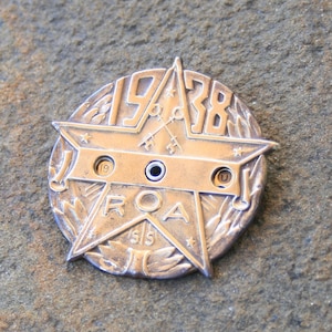 Può includere: Un emblema in metallo color argento a forma di stella con il testo "1938" e "ROAD" attorno alla stella. L'emblema ha tre piccoli fori nella parte superiore e inferiore della stella.