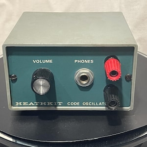 Könnte beinhalten: Ein Vintage Heathkit Code Oszillator in einem grauen Metallgehäuse mit einer teal-farbenen Frontplatte. Zu den Merkmalen gehören ein Lautstärkeregler, eine Kopfhörerbuchse und rote und schwarze Anschlüsse. Der Text "VOLUME", "PHONES" und "HEATHKIT CODE OSCILLATOR" ist sichtbar.