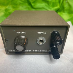 Puede incluir: Un dispositivo electrónico verde oliva vintage con las palabras "VOLUME" y "PHONES" en el panel frontal. Cuenta con una perilla de volumen, un conector para auriculares y dos perillas negras. También es visible el texto "HE CIT CODE OSCILLATOR".