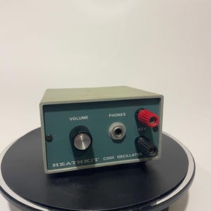 May include: A vintage Heathkit Code Oscillator. The device has a teal front panel with controls for volume, phones, and key. It features red and black terminals and a silver knob. The text "Heathkit Code Oscillator" is printed on the front.