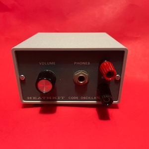 Op de afbeelding: Een vintage Heathkit Code Oscillator, een grijs rechthoekig apparaat met een donkergrijs voorpaneel. Het heeft een volumeknop, een koptelefoonaansluiting en rode en zwarte aansluitingen. De tekst "VOLUME" en "PHONES" is zichtbaar.
