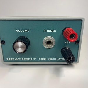 May include: A Heathkit Code Oscillator in a gray metal housing with a teal front panel. The panel features a black volume knob, a central headphone jack labeled PHONES, and two binding posts labeled KEY, one red and one black, with white text.