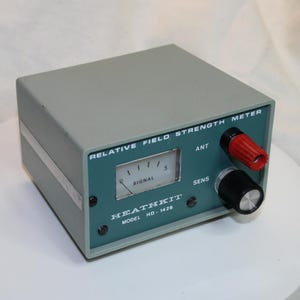 Puede incluir: Un medidor de intensidad de campo relativo Heathkit modelo HD-1426 gris con panel frontal azul verdoso. El dispositivo cuenta con un medidor de señal analógico, un terminal de antena rojo etiquetado como ANT y un mando de sensibilidad negro.