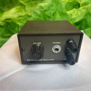 Puede incluir: Un oscilador de código Heathkit gris oscuro. El panel frontal tiene una perilla de volumen, un conector para auriculares y dos perillas negras etiquetadas "KE". El texto "Heathkit Code Oscillator" está impreso debajo de la perilla de volumen.