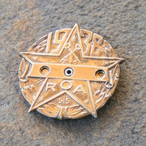 Può includere: Un distintivo a forma di stella in metallo dorato con il testo "ROA" e "1928" su di esso. Il distintivo ha tre fori nella parte superiore e il testo "SIS" nella parte inferiore.