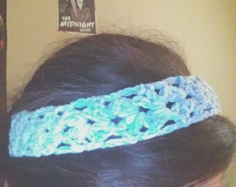 May include: A blue and green crocheted headband with a star pattern. The headband is worn by a person with long dark hair.
