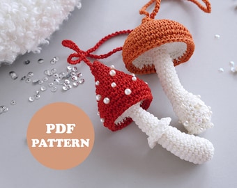 May include: Two crocheted mushroom ornaments, one red with white dots and one orange, with white stems. The mushrooms are hanging from a white fluffy surface. The text "PDF PATTERN" is visible in the image.
