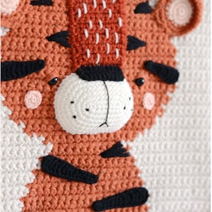 May include: A crocheted tiger wall hanging with a white background and orange and black details. The tiger has a white nose and black eyes.