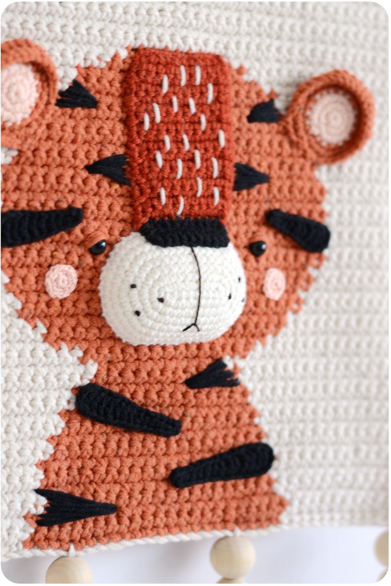 May include: A crocheted tiger wall hanging with a white background and orange and black details. The tiger has a white nose and black eyes.