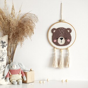 May include: A brown bear face is needle-punched on a white canvas and framed in a wooden hoop. The hoop is hanging from a white wall with three white tassels hanging from the bottom.