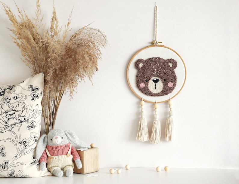 May include: A brown bear face is needle-punched on a white canvas and framed in a wooden hoop. The hoop is hanging from a white wall with three white tassels hanging from the bottom.