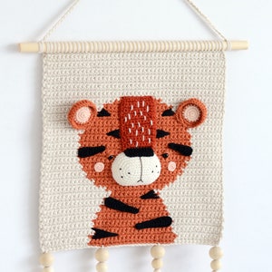May include: A crocheted wall hanging featuring a tiger with orange and black stripes. The tiger has a beige background and wooden beads with tassels hanging from the bottom.