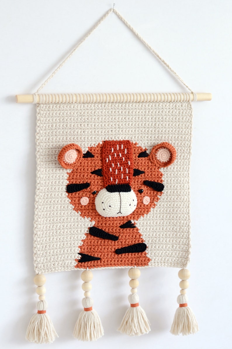 May include: A crocheted wall hanging featuring a tiger with orange and black stripes. The tiger has a beige background and wooden beads with tassels hanging from the bottom.