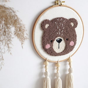 May include: A brown bear face is needle-punched on a white canvas in a wooden embroidery hoop. The hoop is hanging from a white wall with three white tassels hanging from the bottom.
