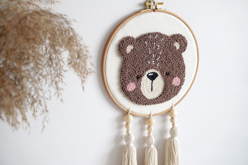 May include: A brown bear face is needle-punched on a white canvas in a wooden embroidery hoop. The hoop is hanging from a white wall with three white tassels hanging from the bottom.