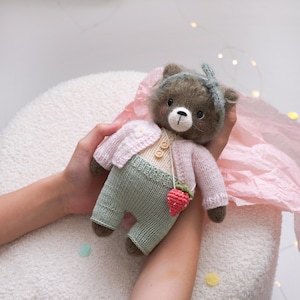 May include: A handmade brown teddy bear doll wearing a light pink cardigan, green pants, and a strawberry purse. The bear has a blue headband and a white nose. The doll is made of soft, textured yarn.