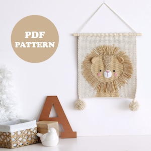 May include: A crocheted lion wall hanging with a beige background and brown yarn. The lion has a pink nose and eyes. The hanging has two beige pom-poms on the bottom. The text "PDF PATTERN" is in a circle on the left side of the image.