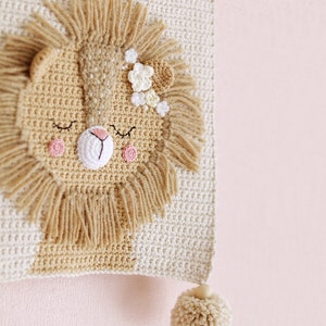 May include: A crocheted lion head wall hanging with a beige background and a brown mane. The lion has pink cheeks and a flower crown. The hanging has a beige pom-pom at the bottom.