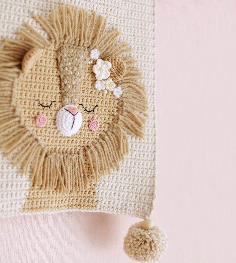 May include: A crocheted lion head wall hanging with a beige background and a brown mane. The lion has pink cheeks and a flower crown. The hanging has a beige pom-pom at the bottom.