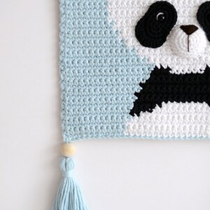 May include: A light blue crocheted wall hanging with a panda face design. The panda has a black nose, black eyes, and a black and white face. The panda is wearing a flower crown with pink and white flowers. The wall hanging has a light blue tassel with a wooden bead at the top.