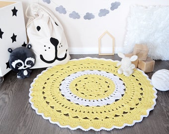 May include: A yellow and white crocheted round rug with a white border. The rug is on a wooden floor with a stuffed animal and a white pillow.