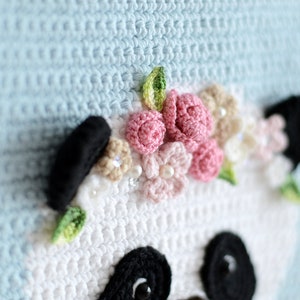 May include: Crocheted panda bear with a pink and white flower crown. The panda is white with black eyes and ears. The background is light blue.