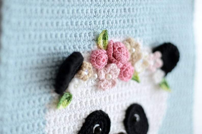 May include: Crocheted panda bear with a pink and white flower crown. The panda is white with black eyes and ears. The background is light blue.