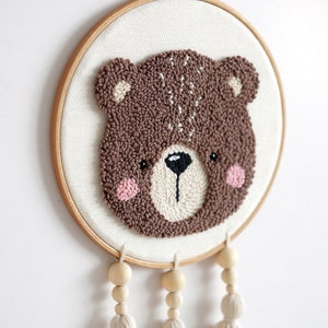 May include: A brown bear face is embroidered on a white canvas in a wooden embroidery hoop. Three wooden beads with white tassels hang from the bottom of the hoop.