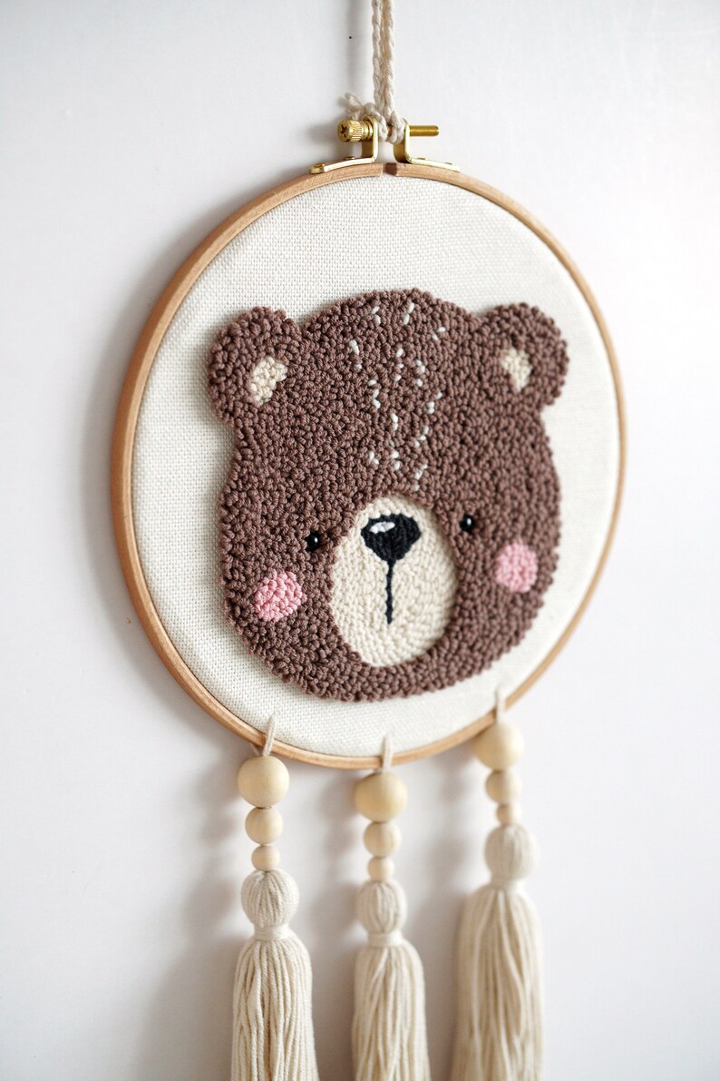 May include: A brown bear face is embroidered on a white canvas in a wooden embroidery hoop. Three wooden beads with white tassels hang from the bottom of the hoop.
