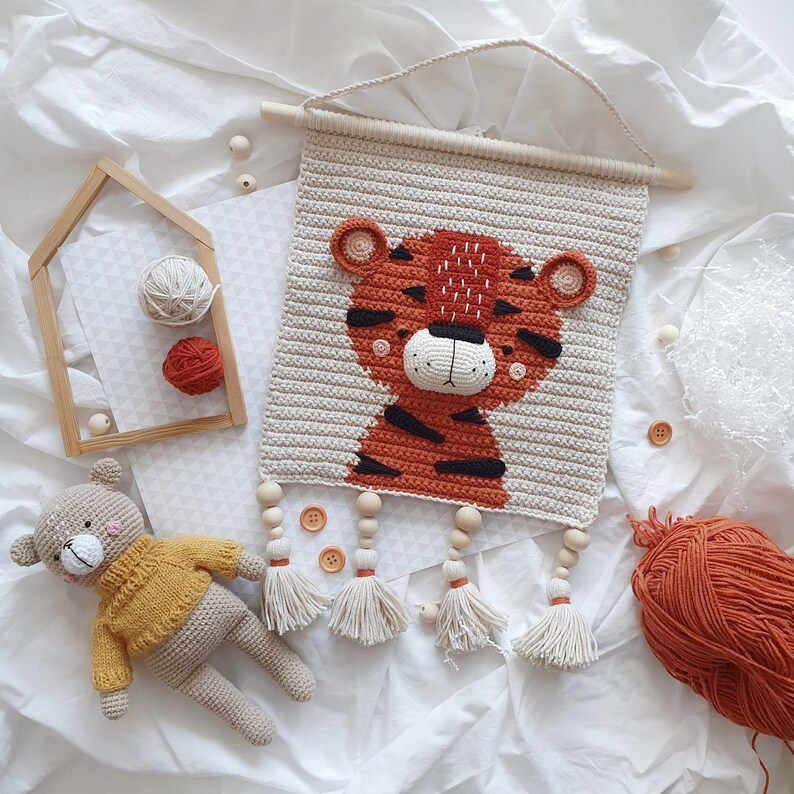 May include: A crocheted wall hanging featuring a tiger with orange and black stripes. The tiger has a white face with black eyes and a pink nose. The wall hanging is decorated with wooden beads and tassels.