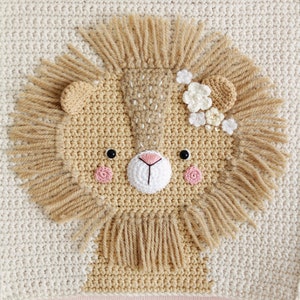 May include: A crocheted lion head wall hanging with a beige background and a brown lion face with a white flower crown. The lion has pink cheeks and black eyes.