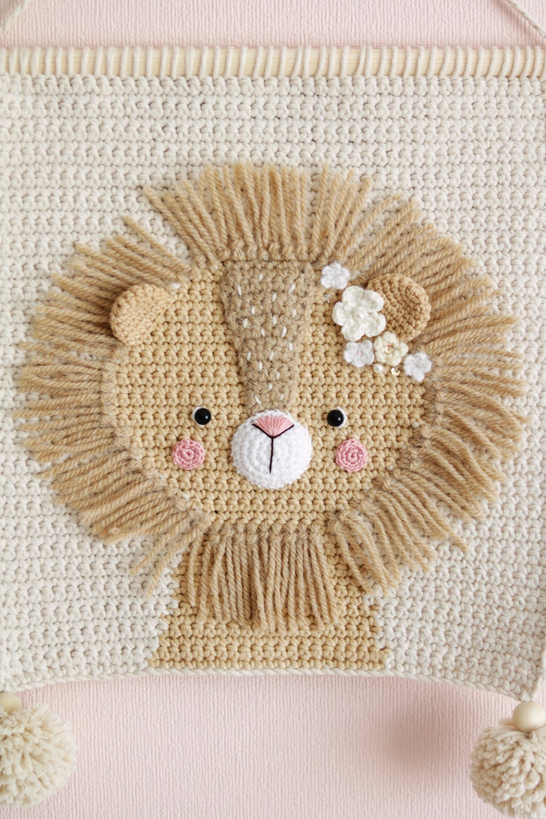 May include: A crocheted lion head wall hanging with a beige background and a brown lion face with a white flower crown. The lion has pink cheeks and black eyes.