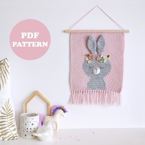 May include: A crochet wall hanging featuring a grey bunny with pink ears and a flower crown. The bunny has closed eyes and a pink nose. The wall hanging is made with pink yarn and has a fringe at the bottom. The text "PDF PATTERN" is written in white on a pink circle.
