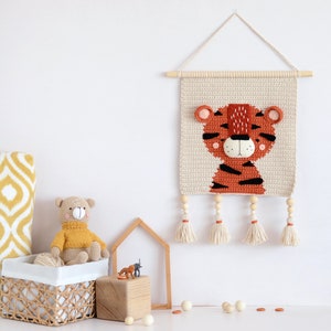 May include: A crocheted tiger wall hanging with a beige background and orange and black stripes. The tiger has a white face with black eyes and a pink nose. The hanging has wooden dowels and tassels.