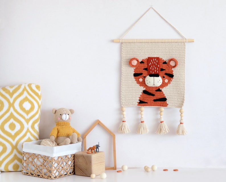 May include: A crocheted tiger wall hanging with a beige background and orange and black stripes. The tiger has a white face with black eyes and a pink nose. The hanging has wooden dowels and tassels.