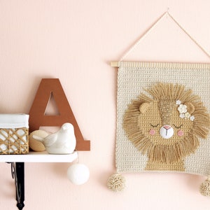 May include: A crocheted wall hanging featuring a lion with closed eyes and pink cheeks. The lion has a brown mane and is decorated with white flowers. The wall hanging is beige and has two white pom-poms at the bottom.