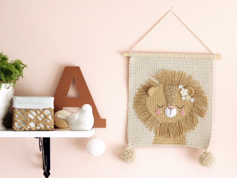 May include: A crocheted wall hanging featuring a lion with closed eyes and pink cheeks. The lion has a brown mane and is decorated with white flowers. The wall hanging is beige and has two white pom-poms at the bottom.