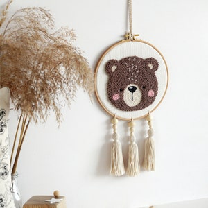 May include: A brown bear face is needle-punched on a white canvas in a wooden embroidery hoop. The hoop is hanging from a white cord and has three white tassels hanging from it.