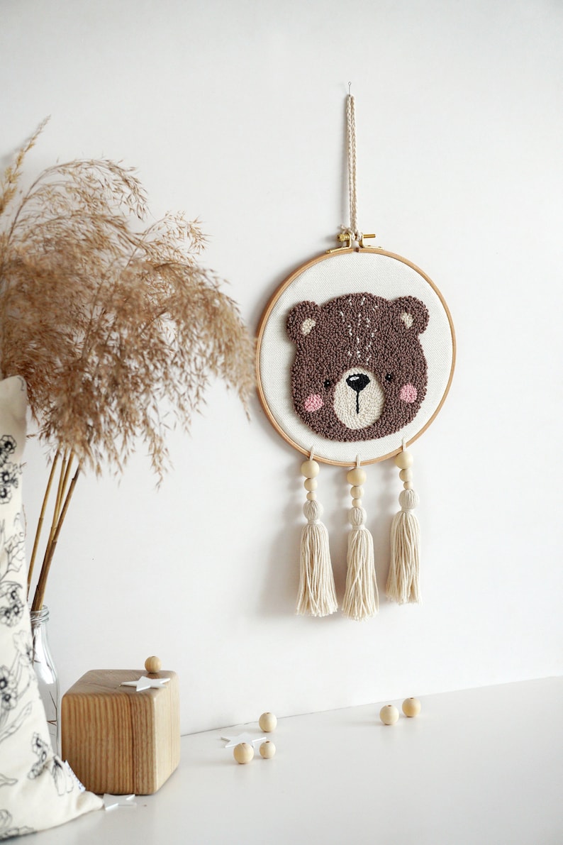 May include: A brown bear face is needle-punched on a white canvas in a wooden embroidery hoop. The hoop is hanging from a white cord and has three white tassels hanging from it.