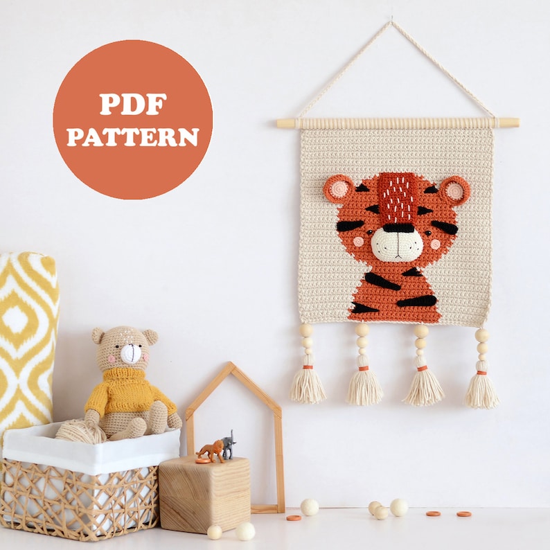 May include: Crochet pattern for a tiger wall hanging with wooden beads and tassels. The pattern is available as a PDF download. The image also shows a crocheted tiger plushie and a wooden house.