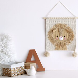 May include: A crocheted wall hanging featuring a lion's face with a beige background and brown yarn for the mane. The hanging has two beige pom-poms on the bottom corners.
