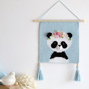 May include: A crocheted wall hanging featuring a panda with a flower crown. The panda is white with black ears, eyes, nose, and a black bow tie. The background is light blue and the hanging has two blue tassels.
