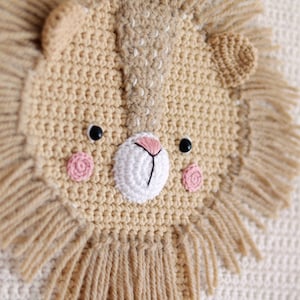 May include: A crocheted lion head wall hanging with a beige face and pink cheeks. The lion has a white crocheted nose and black eyes. The mane is made of beige yarn with long strands.