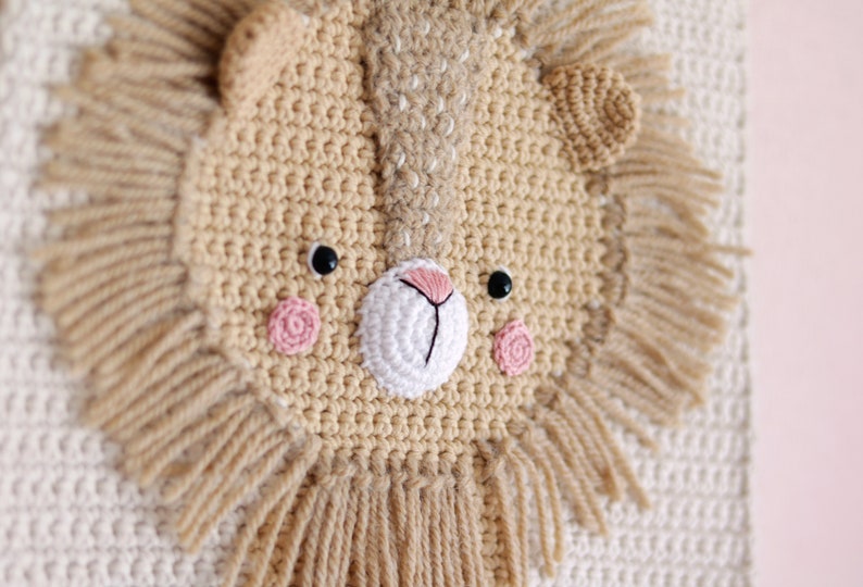 May include: A crocheted lion head wall hanging with a beige face and pink cheeks. The lion has a white crocheted nose and black eyes. The mane is made of beige yarn with long strands.