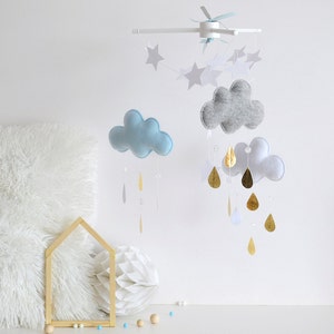 Puede incluir: Un móvil para bebé con nubes de fieltro gris, blanco y azul, gotas de lluvia de lámina dorada y estrellas blancas. El móvil está colgado de un marco de metal blanco.