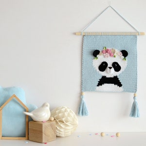 May include: A blue crocheted wall hanging featuring a panda with a flower crown. The panda is white with black ears, eyes, and a nose. The hanging has light blue tassels on each side.
