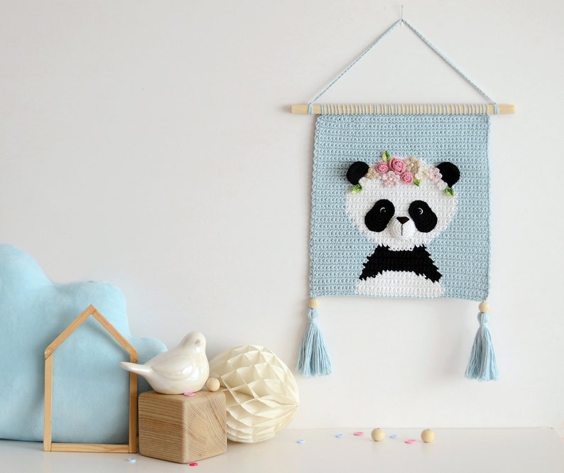 May include: A blue crocheted wall hanging featuring a panda with a flower crown. The panda is white with black ears, eyes, and a nose. The hanging has light blue tassels on each side.