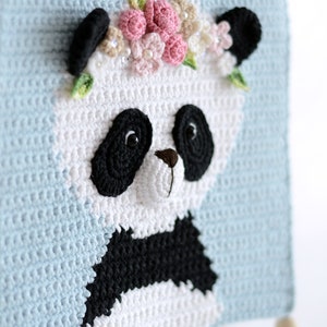 May include: A crocheted panda bear with black eyes and a black nose, wearing a pink and white flower crown. The panda is on a light blue background with a white tassel.
