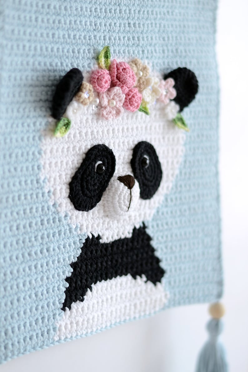 May include: A crocheted panda bear with black eyes and a black nose, wearing a pink and white flower crown. The panda is on a light blue background with a white tassel.