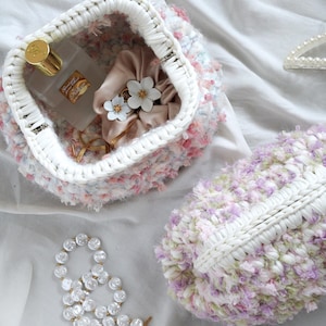 Puede incluir: Un bolso y un clutch a juego hechos a mano y esponjosos en colores pastel, con un ribete blanco tejido. El bolso contiene un frasco de perfume, una goma para el pelo con detalles de flores y joyas doradas. Un collar de perlas y pequeñas flores completan la escena.